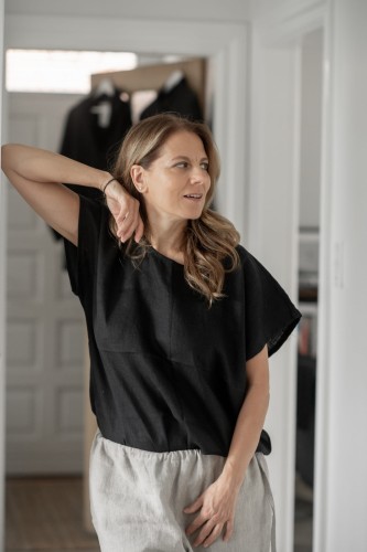 Uśmiechnięta kobieta stoi w jasnym pokoju. Ma na sobie luźną, czarną koszulkę lnianą oraz beżowe spodnie z lnu.