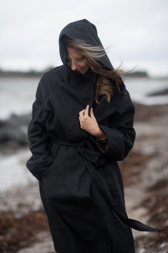 Kobieta spaceruje po plaży. Ma na sobie długi, czarny płaszcz z lnu. Płaszcz posiada kieszenie po bokach i jest wiązany po środku lniany paskiem.