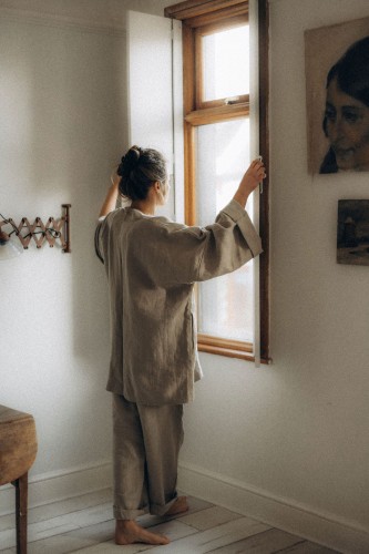 Kobieta patrzy przez okno. Jest ubrana w beżowe kimono lniane z podwiniętymi rękawami oraz bezowe spodnie z lnu.