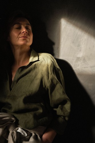 Uśmiechnięta kobieta stoi oparta o czarną ścianę. Ma na sobie zieloną koszulę z lnu o luźnym kroju. Na biodrach ma zawiązany szary szalik.