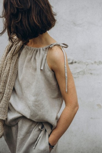 Kobieta stoi przy szarej ścianie. Ma na sobie szary top z lnu o luźnym kroju, szarą sukienkę z lnu oraz beżowy szal.
