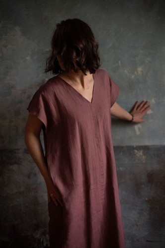 Kobieta opiera się ręką o czarną ścianę. Jest ubrana w luźny, bordowy kaftan z lnu.